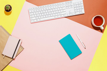Modern home office workspace. Flat lay composition of keyboard, cactus, diary, notebook with pen and cup of tea on colorful desk. Pink, yellow, aquamarine and and brown colours