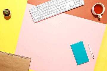 Trendy home office workspace. Flat lay composition of keyboard, cactus, diary with pen and cup of tea on colorful desk. Pink, yellow, aquamarine and and brown colours. Blogger's home office