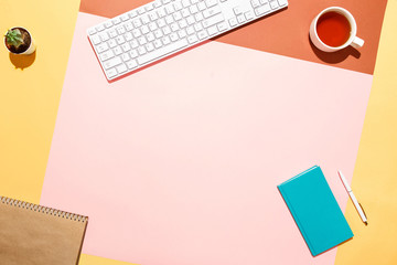 Trendy home office workspace. Flat lay composition of keyboard, cactus, diary with pen and cup of tea on colorful desk. Pink, yellow, aquamarine and and brown colours. Blogger's home office