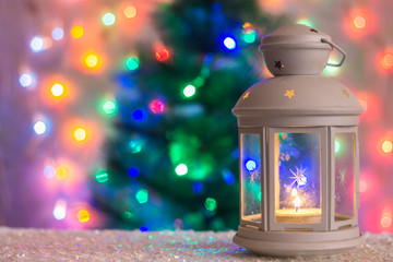 Christmas white lantern with a burning candle on the background of a Christmas tree, lights of garlands, bokeh.
