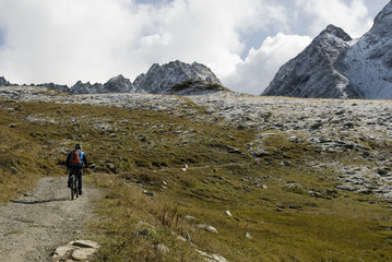 Fototapeta premium man with an ebike, electric bike, mountains covered with fresh snow just fall, autumn, observes Kastelhorn (Castel Peak), grass, dirt, October, Alps, light, clouds, Formazza valley, Piedmont, Italy