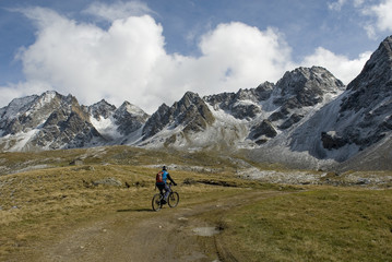 Fototapeta na wymiar man with an ebike, electric bike, mountains covered with fresh snow just fall, autumn, observes Kastelhorn (Castel Peak), grass, dirt, October, Alps, light, clouds, Formazza valley, Piedmont, Italy
