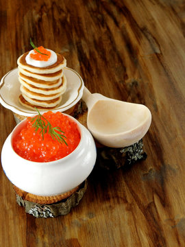 Red caviar in a white ceramic bowl and pancakes with caviar on a wooden table