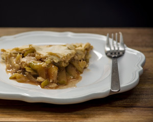 A slice of apple pie on white plate on wooden table, viewed from the side, decorated with a fork, against black wall