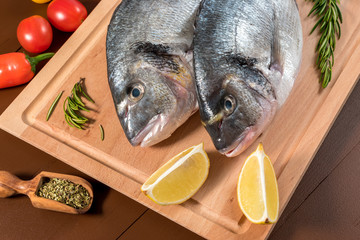 Fresh uncooked dorado or sea bream fish with lemon, herbs, vegetables and spices on wooden board over dark backdrop, top view.