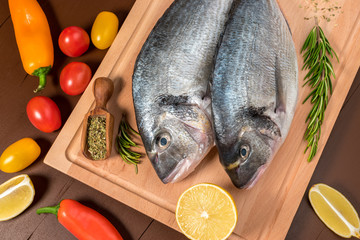 Fresh uncooked dorado or sea bream fish with lemon, herbs, vegetables and spices on wooden board over dark backdrop, top view.