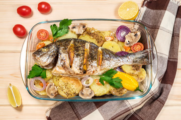 A delicious whole baked fish. Baked sea bream with lemon, onions, herbs, cherry tomatoes, spices and baked potatoes on a wooden background. Fried tasty fish. Diet and healthy food. Top view.