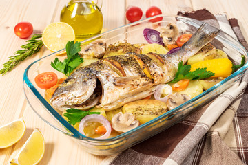 A delicious whole baked fish. Baked sea bream with lemon, onions, herbs, cherry tomatoes, spices and baked potatoes on a wooden background. Fried tasty fish. Diet and healthy food. Top view.