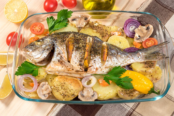 A delicious whole baked fish. Baked sea bream with lemon, onions, herbs, cherry tomatoes, spices and baked potatoes on a wooden background. Fried tasty fish. Diet and healthy food. Top view.