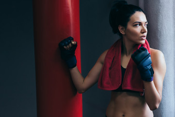 Horizontal shot of strong attractive brunette woman with red towel on neck after workout with punch bag, wearing kickboxing gloves in the gym. Sport, fitness, lifestyle and people concept.
