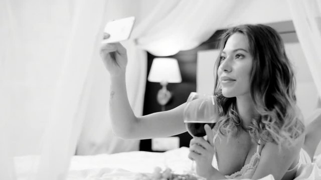 Attractive woman drinks red wine and takes photo of herself on her smartphone on bed in black and white