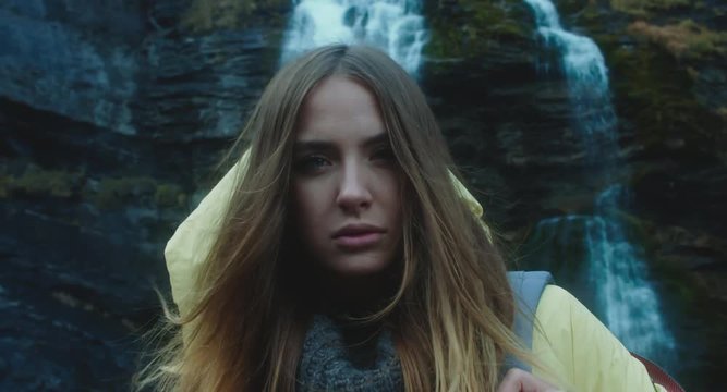 CU Portrait of attractive female hiker wearing yellow raincoat standing on mountain point with a waterfall in the background. 4K UHD RAW edited footage
