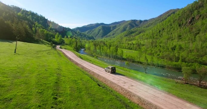 UHD 4K aerial of vehicle driving towards the sunrice. Mid-air shot around a dirty suv retro car driving on green countryside rural road.