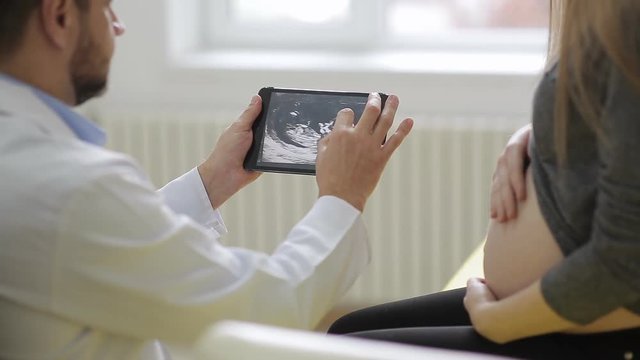 male doctor in medical robe shows a pregnant woman a picture of an ultrasound on a tablet