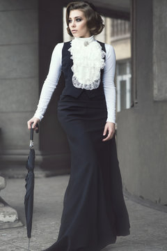 Portrait of gorgeous woman with elegant Victorian hairstyle wearing old-fashioned gown with jabot walking among the pillars of her old castle leaning on black umbrella. Vintage style. Outdoor shot