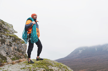 Hipster-climber stands in a down jacket on a rock with a rope on his shoulder