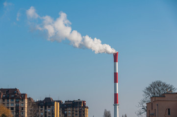 Obraz premium Red and white industrial chimney smoking with blue sky and residential buildings in the background