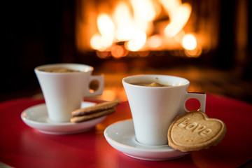 Two coffee cups with heart shaped biscuits on red table with fireplace in background