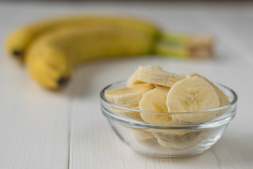 Two whole ripe banana and a glass bowl with slices on a white table.