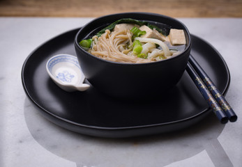 Japanese udon noodle soup with enoki mushroom, tofu and Gai lan. Black ceramic bowl and a plate. Horizontal layout. 
