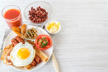 Full English breakfast with smoked sausages, scrambled eggs, bacon, tomatoes, mushrooms, toast and beans. A glass of fresh juice. Breakfast on a white plate on a light background. Space for text. 