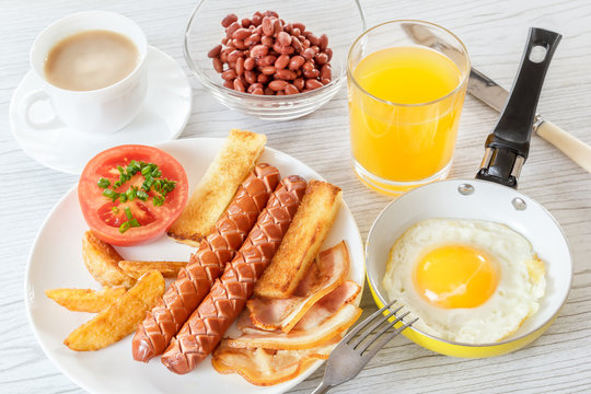 Traditional English breakfast with smoked sausages, bacon, tomato, toast and beans. Fried egg in a frying pan. Tea with milk. A glass of fresh juice. Backlight