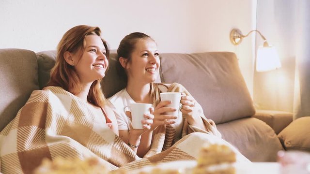 Two young women in plaide are watching TV and warming with tea at home.
