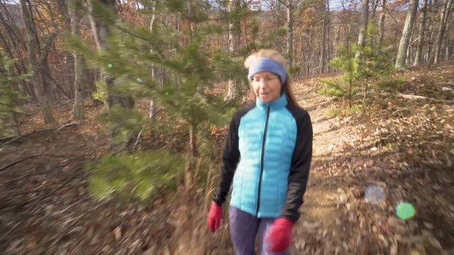 Side closeup shot with lens flares of mature woman wearing blue jacket, headband and red gloves, hiking in the forest with a carpet of autumn leaves and labrador retriever dog.