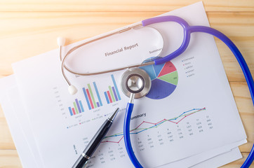 Top view of wooden working table with financial report, stethoscope and pen. Business Concept.