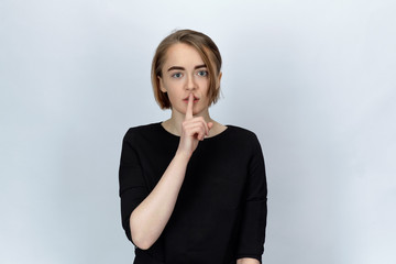 Studio portrait on isolated background of a young attractive woman with wide open eyes, holding a finger to call for silence