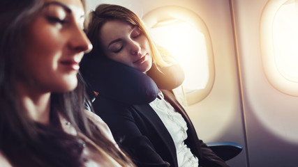 Girlfriends traveling by plane. A female passenger sleeping on neck cushion in airplane. - obrazy, fototapety, plakaty