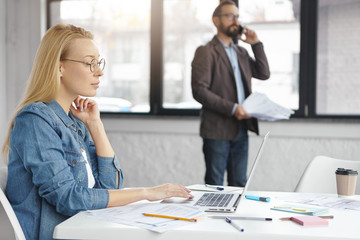 Confident blonde female secretary wears denim shirt and spectacles searches useful information for developing own business in internet website, uses modern laptop while boss has phone conversation