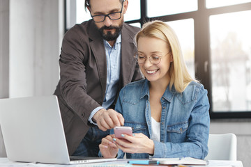 Pretty intelligent female can`t understand how to use new app, asks advice and help in experienced male enterpreneur. Happy business workers prepare for meeting with international partners use gadgets