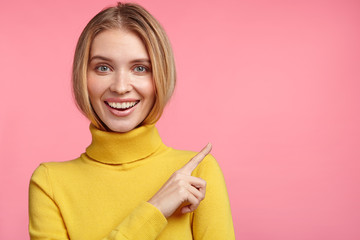 Positive good looking woman has healthy skin, pleasant smile and appealing appearance, points at...