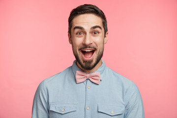 Emotive bearded man wears formal shirt and bow tie, happy to celebrate anniversary, invites friends on party, glad to recieve presents. Happy young unshaven male in festive clothes has good mood