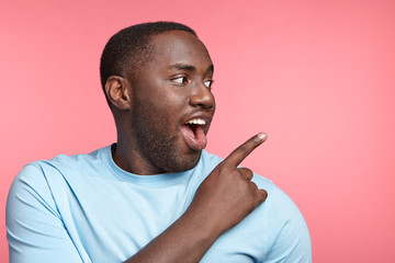Joyful happy black male with excited expression, looks aside, indicates at copy space, sees...