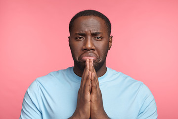 Upset praying sorrorful African male asks for forgiveness, feels guilty, curves lower lip, hopes for apology or excuse, isolated over pink background. Young dark skinned male prayer poses indoor