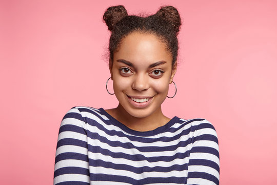 Happy African American young woman with hair knots, wears round earrings, looks directly into camera, to meet international friends, isolated over pink background. People and happiness concept