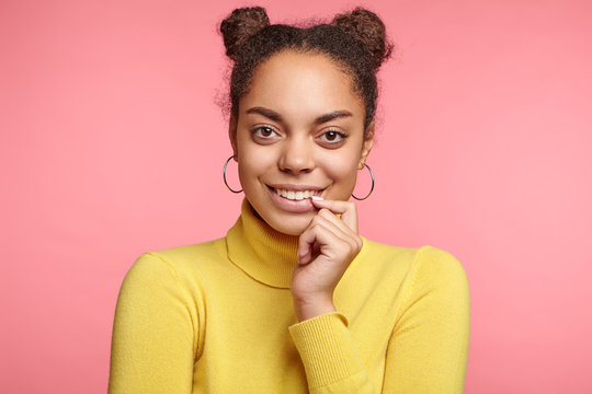 Portrait of happy attractive dark skinned woman with two hair buns, wears yellow polo neck sweater, has pleasant smile, glad to hear compliment about her appearance from handsome guy, isolated on pink