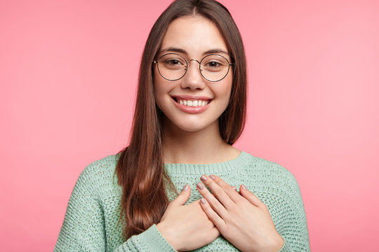 Kind hearted Asian female with long dark hair, wears round glasses, keeps hands on chest, shows her devotion and love to people who surround her, poses against pink background. People and emotions