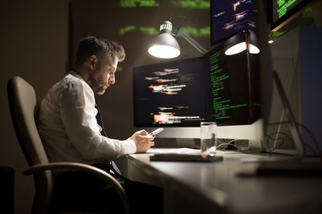 Profile view of talented middle-aged programmer using smartphone while sitting at office desk and...