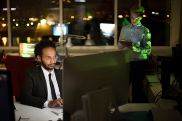 Busy dim open plan office: mixed race manager wearing stylish suit working on promising project with help of computer while his pretty young colleague checking business emails on digital tablet