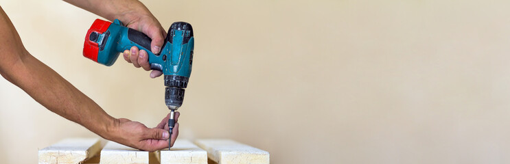 Hand of a worker screws a screw in a wooden board with a cordless screwdriver. Man carpenter at...