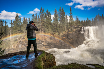 Nature photographer tourist with camera shoots while standing Ristafallet waterfall in Sweden.