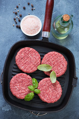 Cast-iron grill pan with raw fresh beef burger cutlets and condiments. High angle view on a blue stone surface