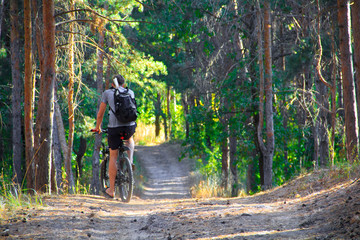 Obraz premium A guy in a gray T-shirt, black shorts and a black backpack is riding along a path in the forest on a bicycle.