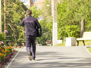 A policeman in uniform with a bag goes to work through the park and talks on his mobile phone.