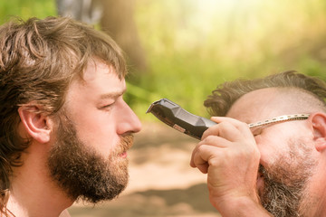 Man with glasses mows beard  the other man using hair clippers