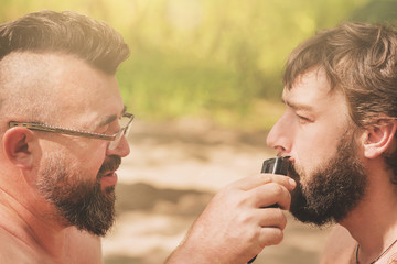 Man with glasses mows beard  the other man using hair clippers
