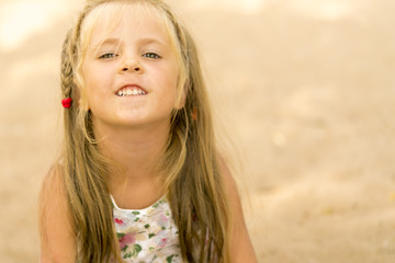 Little blond girl with a pigtail showing teeth on the background of sand